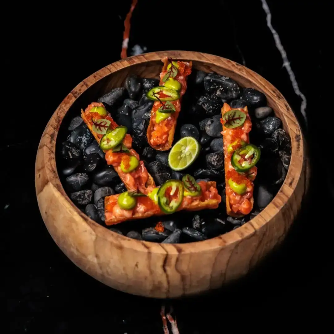 Crispy salmon taquitos standing in a bowl of black stones