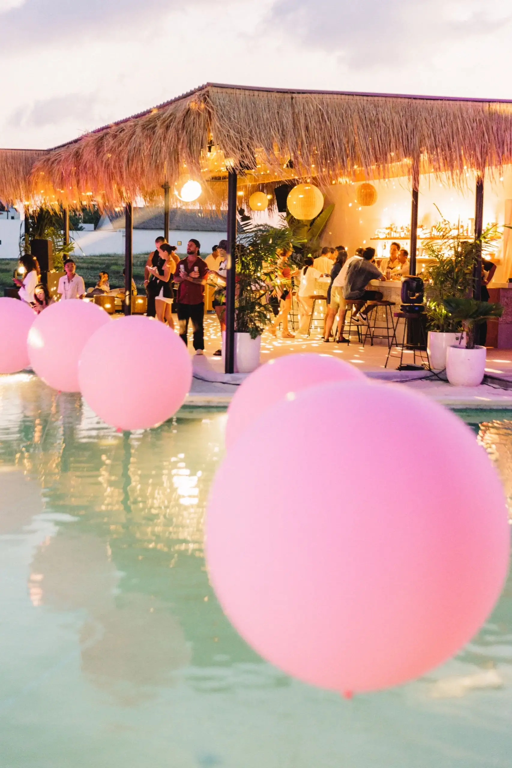 Oversized pink balloons floating on the pool at dusk, the lantern-lit thatch bar behind them