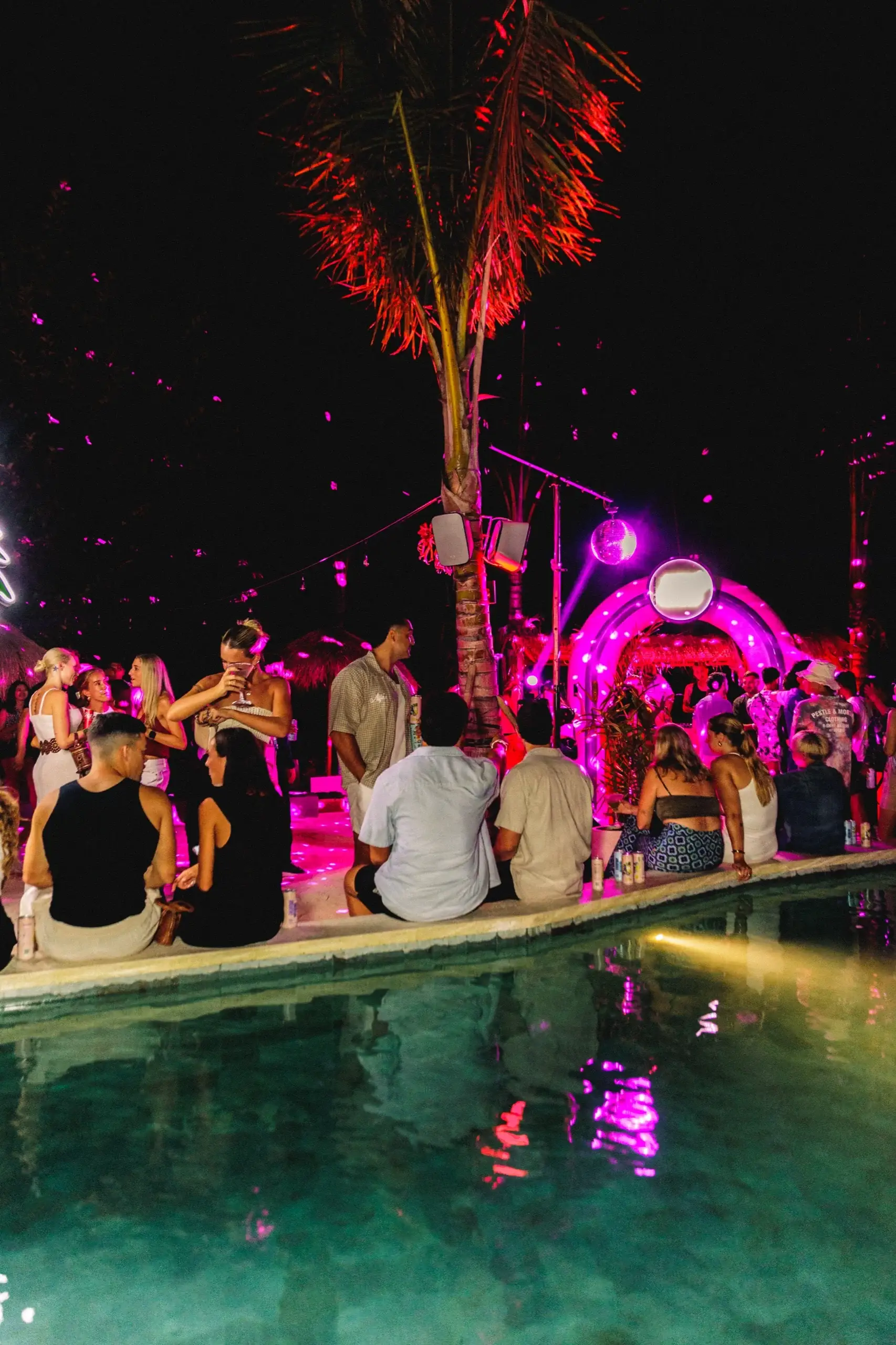 The pool at night with the crowd along its edge, a palm uplit red and the branded arch glowing behind