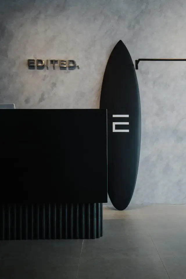 The EDITED. counter in polished concrete, chrome wordmark on the wall behind a black surfboard — EDITED.
