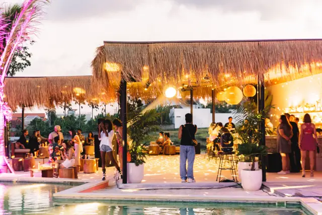 The venue at golden hour — a thatched bar hung with lanterns, the DJ set up on the deck and the pool running along the front — Everyday Weekend